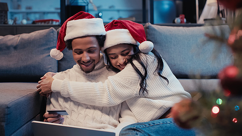 happy african american couple in santa hats hugging while online shopping near blurred christmas tree