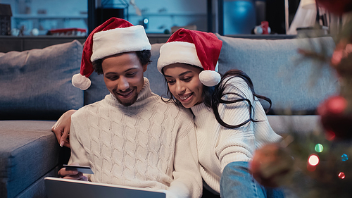 smiling african american couple in santa hats online shopping near blurred christmas tree