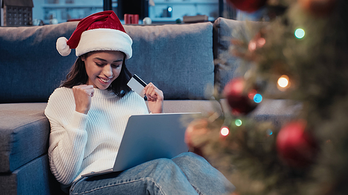 excited african american woman holding credit card and using laptop near christmas tree