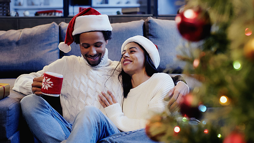 African american man holding cup and smiling with girlfriend near christmas tree