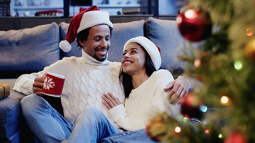 African american man holding cup and smiling with woman near christmas tree