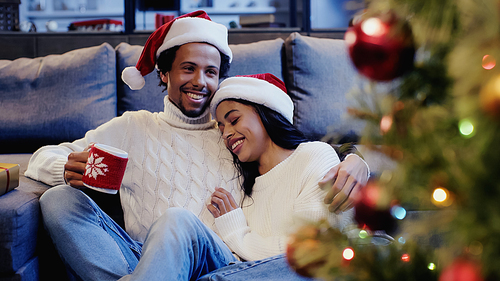 cheerful african american man holding cup and smiling with girlfriend near christmas tree