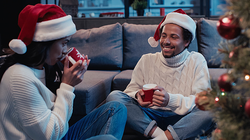 happy african american man in santa hat holding cup and looking at girlfriend drinking near blurred christmas tree