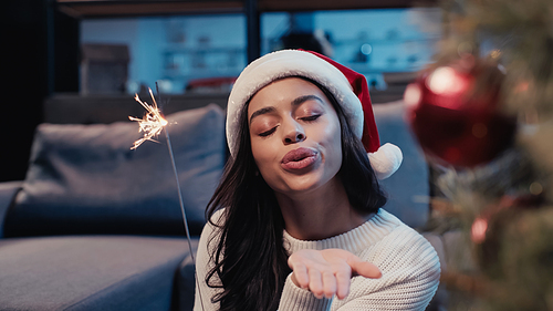 young african american woman in santa hat holding sparkler and sending air kiss near blurred fir