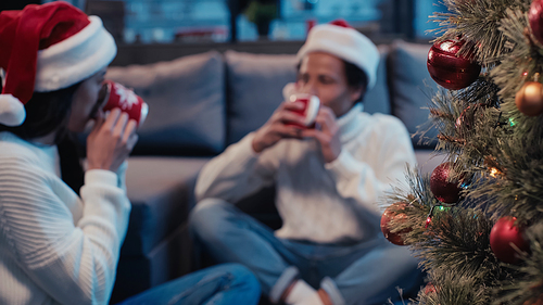 Christmas tree near african american couple holding cups while drinking on blurred background