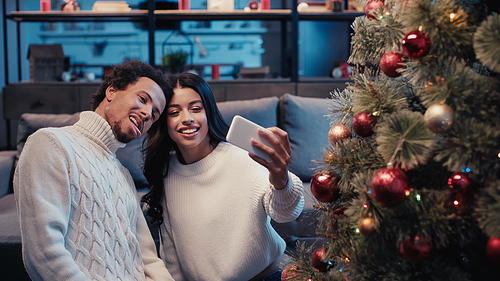 happy african american woman taking selfie with man sticking out tongue near christmas tree