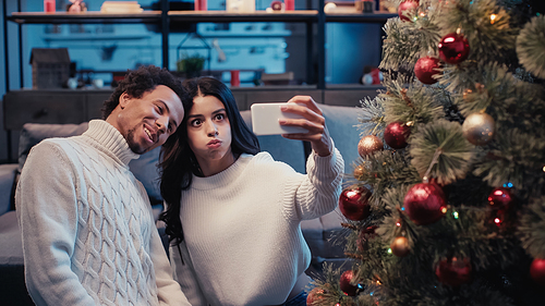 happy african american man taking selfie with woman puffing cheeks near christmas tree