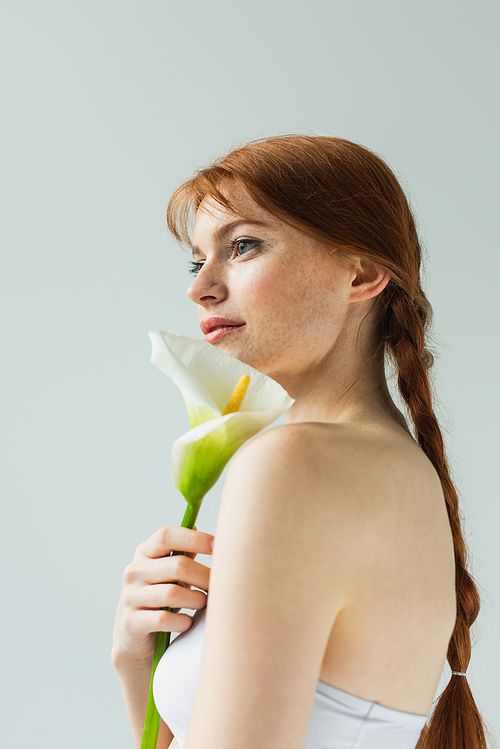 Side view of freckled woman in top holding calla lily isolated on grey