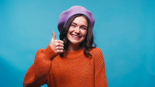 pleased woman in purple beret smiling at camera and showing thumb up isolated on blue