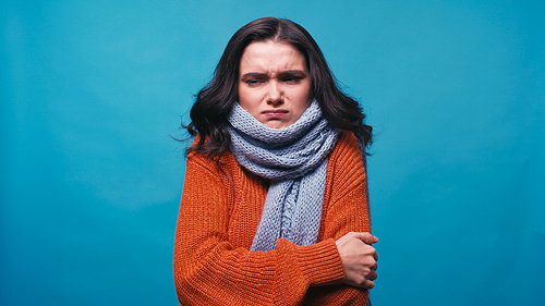 thick and frozen woman in warm scarf hugging herself isolated on blue
