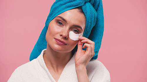 woman with blue towel on head removing eye patch isolated on pink
