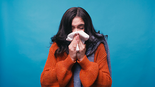 ill woman in warm sweater sneezing in paper napkin isolated on blue