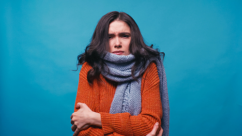 diseased woman in warm scarf and sweater hugging herself isolated on blue