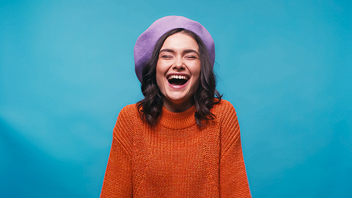 excited woman in beret and jumper laughing with closed eyes isolated on blue