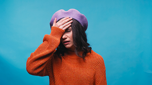 upset woman in beret showing facepalm gesture isolated on blue