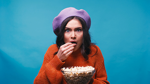 worried woman in purple beret watching film and eating popcorn isolated on blue