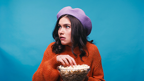 scared woman in beret eating popcorn while watching horror film isolated on blue