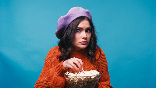 scared woman holding bowl with popcorn while watching horror movie isolated on blue