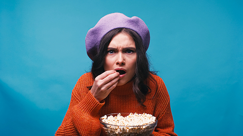 nervous woman eating popcorn while watching exciting film isolated on blue