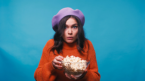 thrilled woman holding bowl with popcorn while watching film isolated on blue