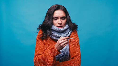 displeased woman in warm scarf and sweater looking at thermometer isolated on blue