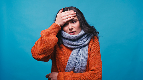 diseased woman in warm sweater and scarf touching forehead isolated on blue