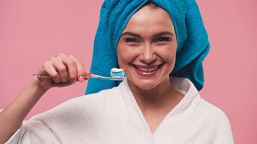 cheerful woman holding toothbrush with toothpaste while smiling at camera isolated on pink
