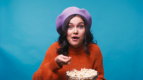 amazed woman in jumper and beret watching exciting movie and eating popcorn isolated on blue