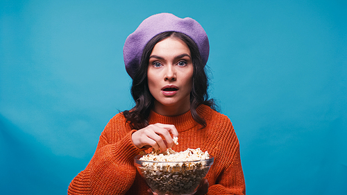 impressed woman holding bowl of popcorn while watching interesting film isolated on blue