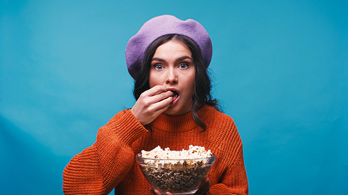 astonished woman in beret watching movie and eating popcorn isolated on blue
