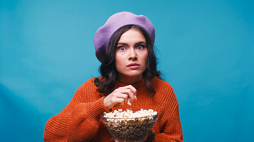 shocked woman holding popcorn while watching movie isolated on blue