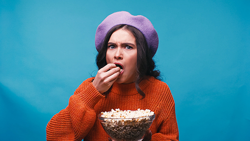 worried woman in lilac beret eating popcorn while watching movie isolated on blue