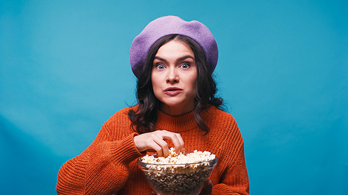 excited woman in sweater and beret holding popcorn while watching film isolated on blue