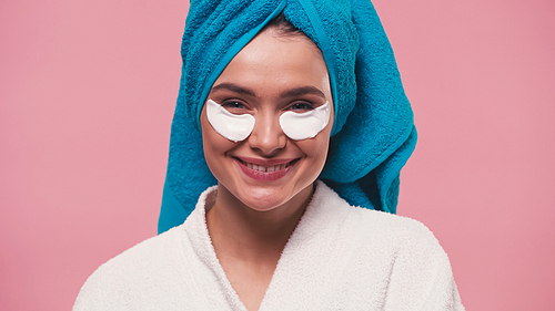 pleased woman with eye patches and towel on head smiling at camera isolated on pink