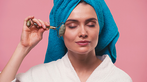 young woman with towel on head massaging face with jade roller isolated on pink