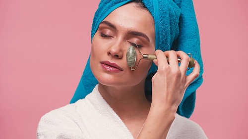 young woman with towel on head and closed eyes massaging face with jade roller isolated on pink