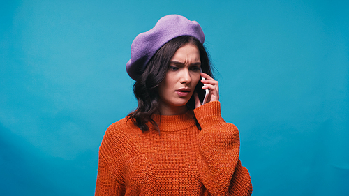thoughtful woman in beret talking on smartphone isolated on blue