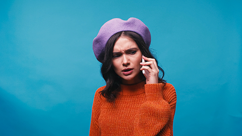 tense woman in purple beret talking on mobile phone isolated on blue