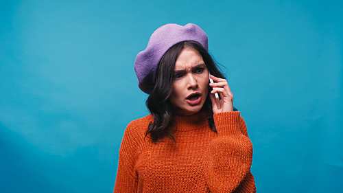 angry woman in sweater and beret talking on mobile phone isolated on blue