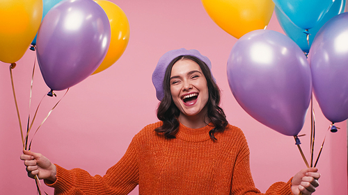 excited woman in beret and jumper laughing near colorful balloons isolated on pink
