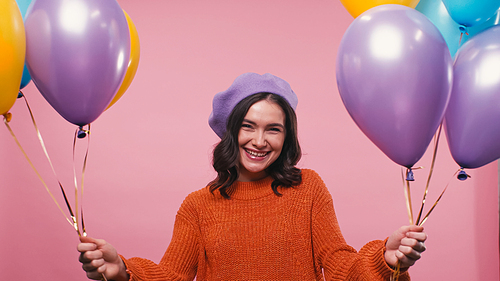 cheerful woman in beret and jumper holding multicolored balloons isolated on pink