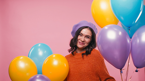 happy woman in beret and sweater smiling near festive balloons isolated on pink