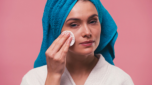 woman with terry towel on head wiping face with cotton pad isolated on pink