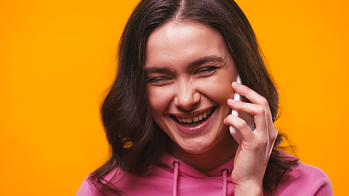 excited brunette woman laughing while talking on cellphone isolated on yellow