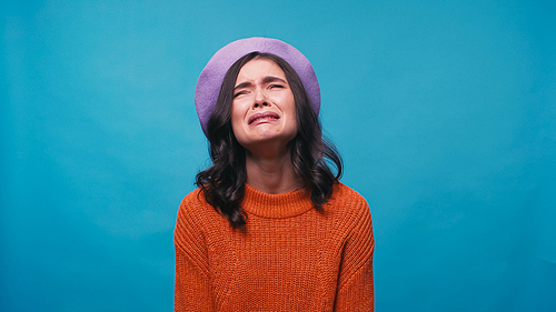 sad woman in warm jumper and beret crying isolated on blue