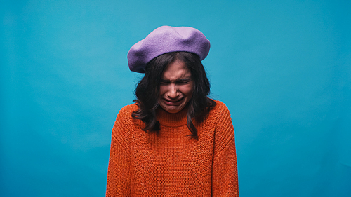 depressed woman in knitted sweater and beret crying isolated on blue