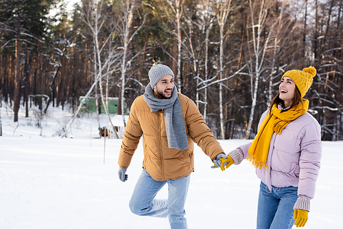 Cheerful man holding hand of excited girlfriend in winter park