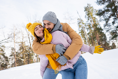 Excited man in knitted hat embracing girlfriend with closed eyes in park with snow