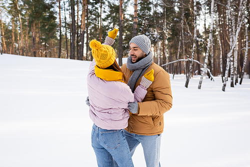 Smiling man looking at girlfriend in knitted hat in park with snow