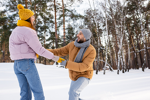Side view of excited man holding hands of girlfriend in gloves in snowy park